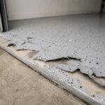 A failed DIY epoxy garage floor kit peeling and lifting at the slab edge, showing flake delamination near the roller door