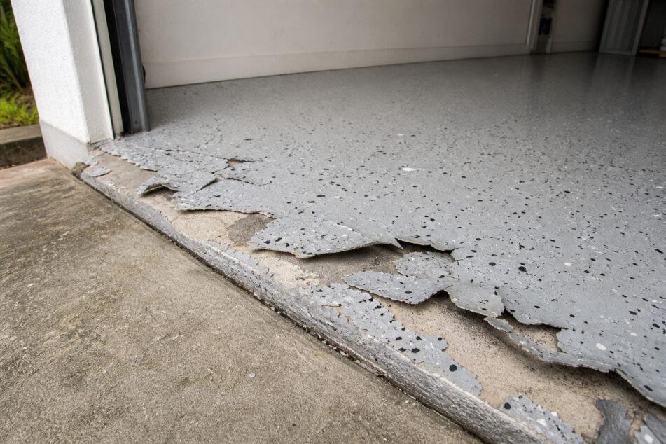 A failed DIY epoxy garage floor kit peeling and lifting at the slab edge, showing flake delamination near the roller door